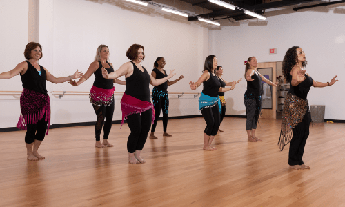 Oreet stands in front of a room full of students wearing hipscarves. She is teaching SharQui, the bellydance workout.